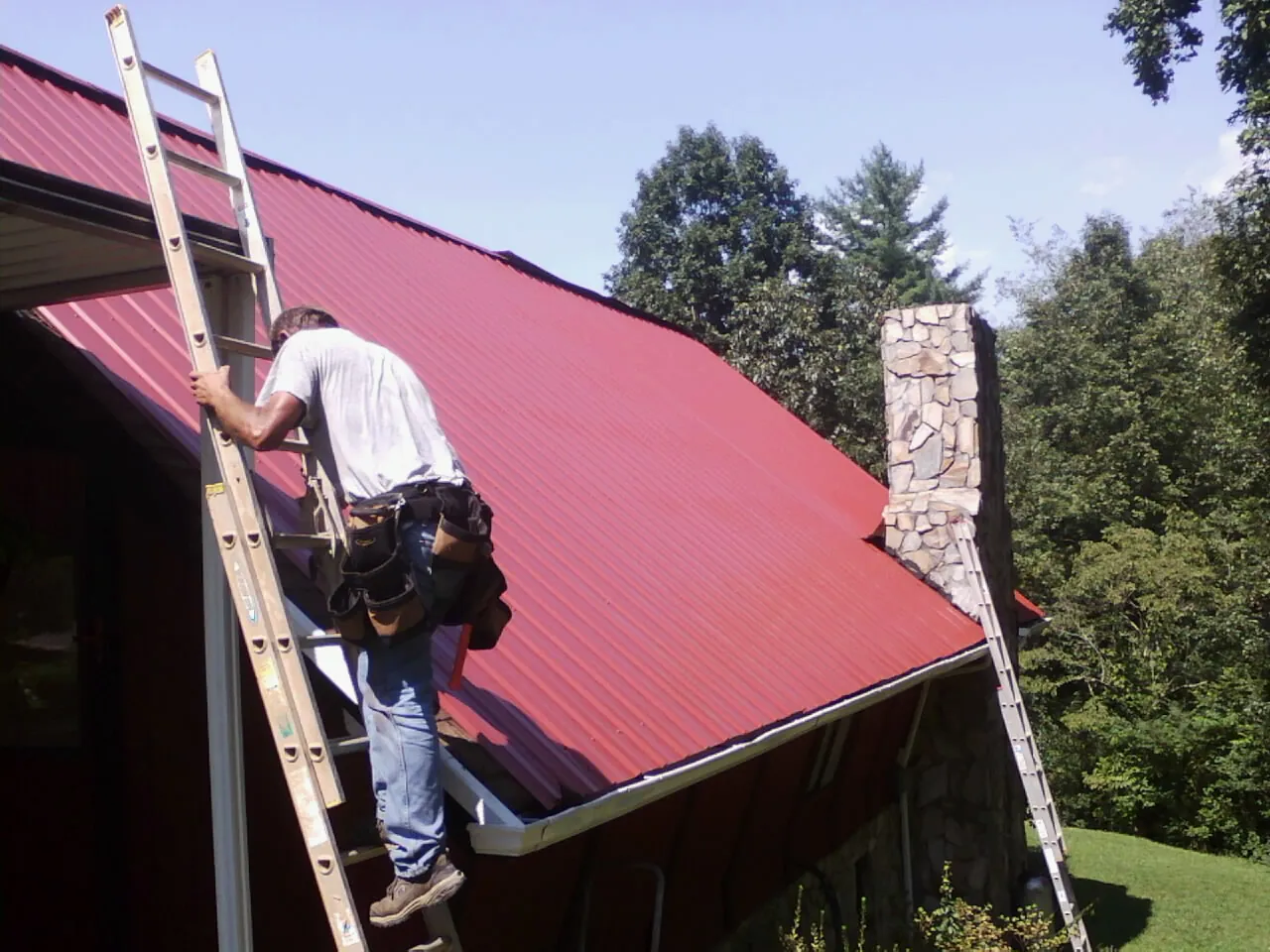 Roofer on a ladder beside a red metal roof and stone chimney — Soffit & Fascia Repair by Roofing Brothers in Lenni